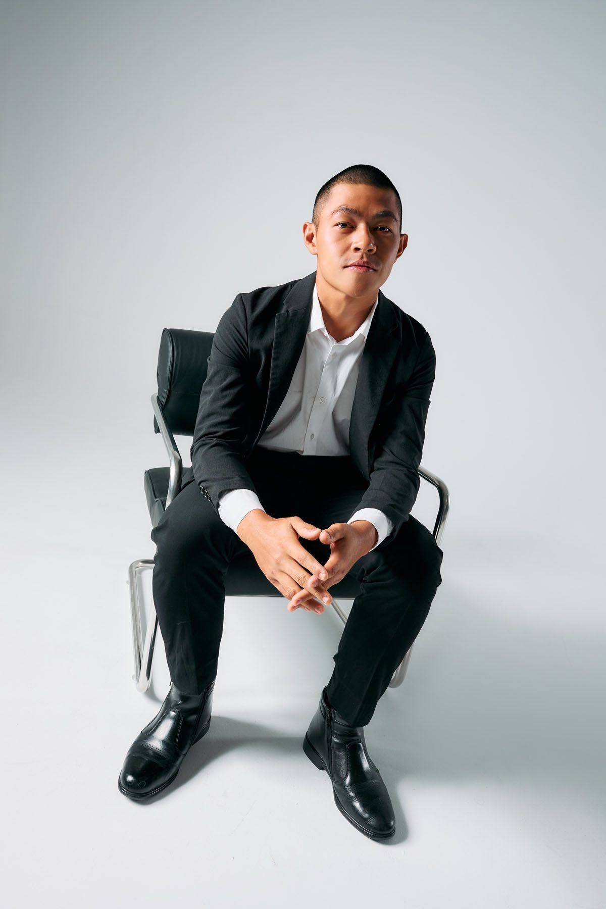 Young Asian man in black suit sitting on chair with hands clasped, posing confidently against gray background.