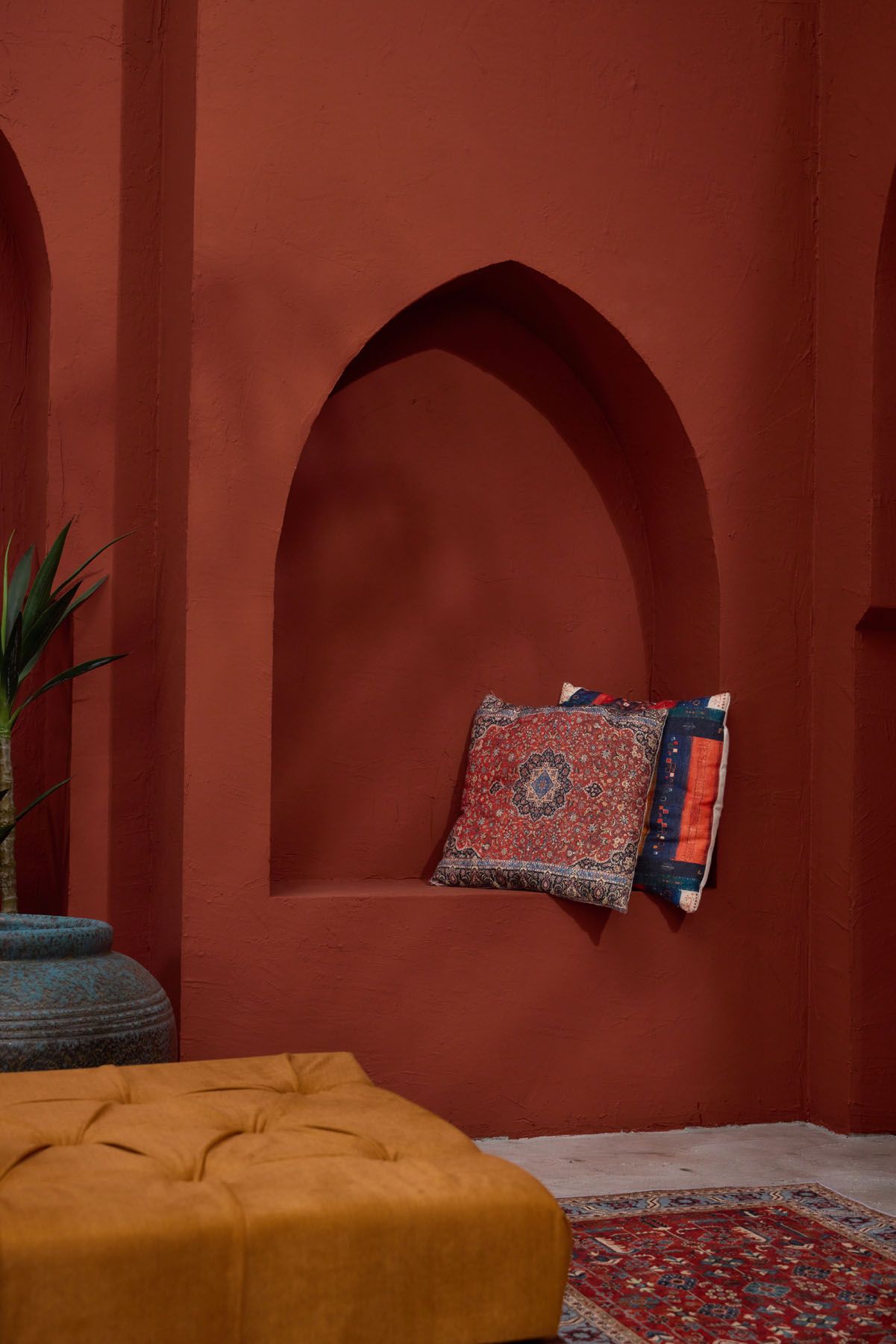 Moroccan-style terracotta red alcove with ornate arch niche displaying patterned cushions above a tufted mustard ottoman.
