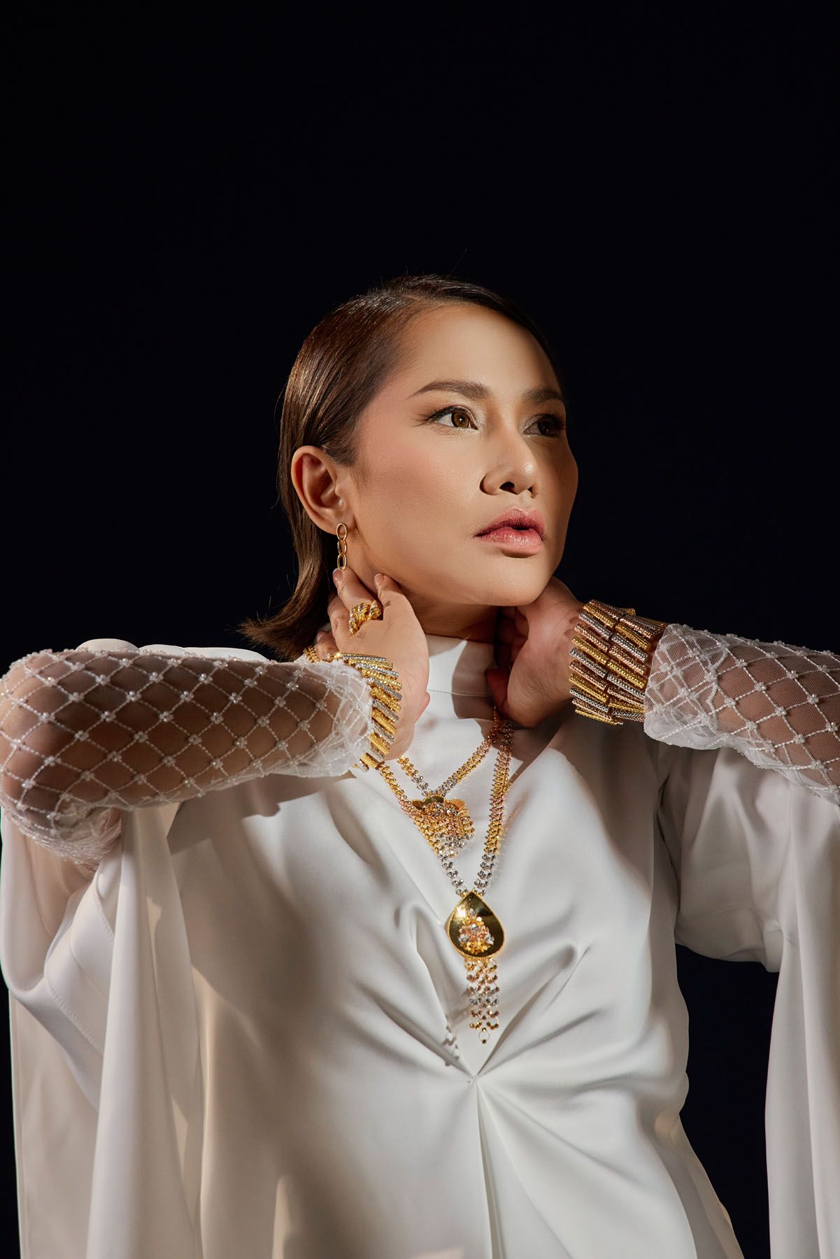 Woman in white outfit with sheer sleeves showcasing gold jewelry necklaces and bracelets on dark background.