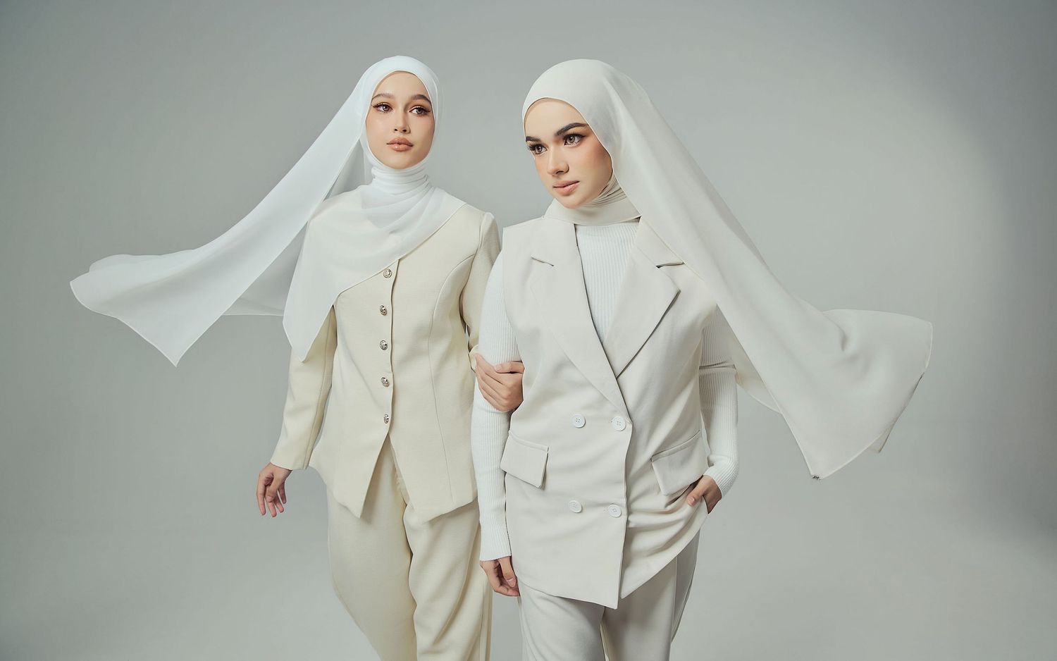 Two women in matching white hijab and tailored white suits posing together in studio setting.