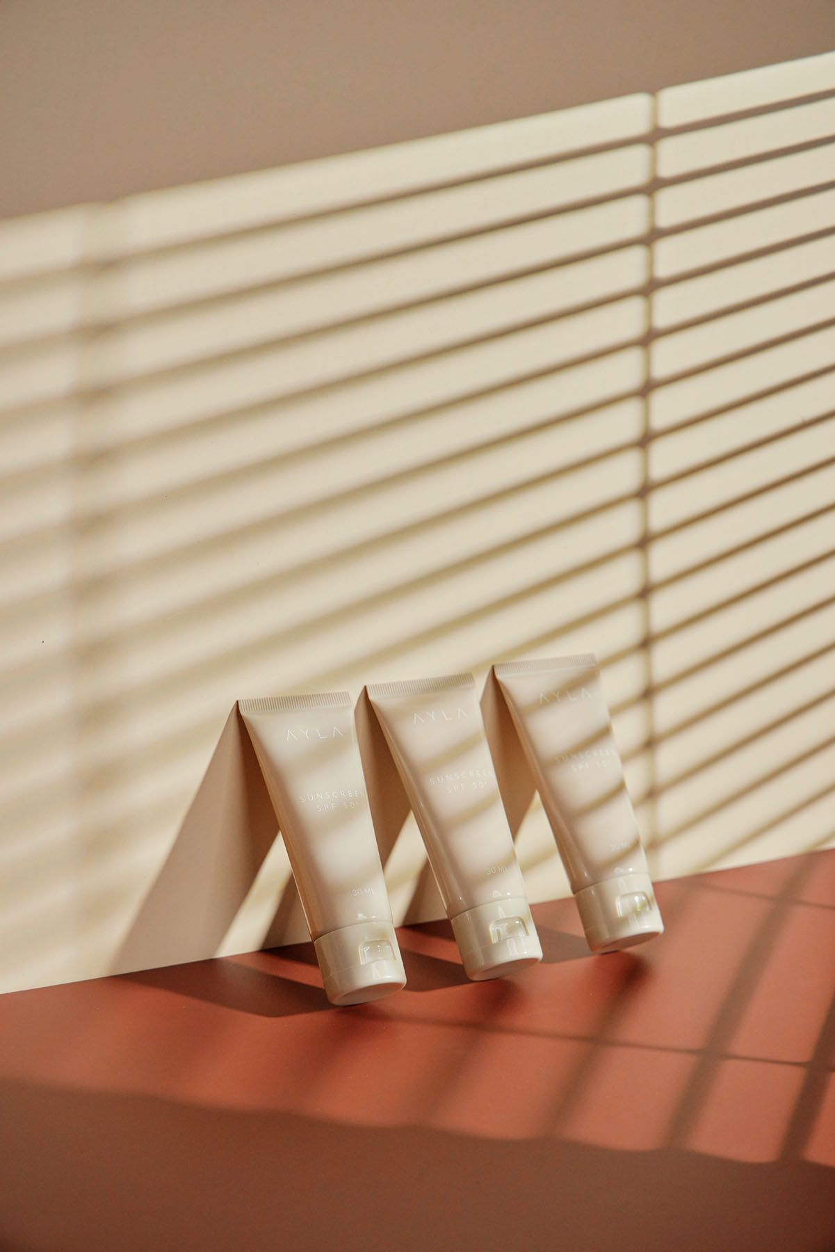 Four beige skincare cream tubes leaning against wall on terracotta surface with striped blind shadow pattern.