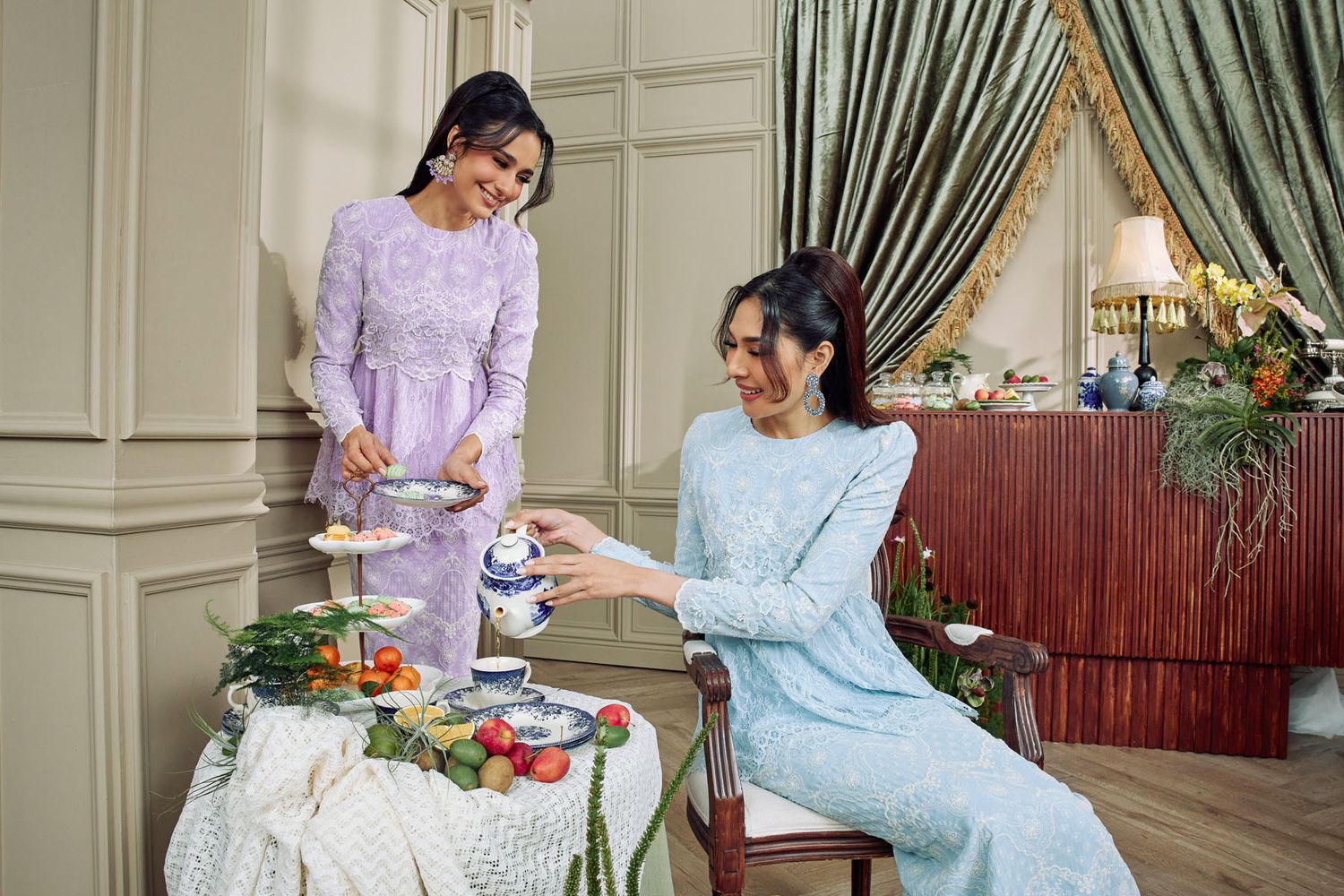 Two Muslim women in pastel baju kurung enjoying afternoon tea in an elegant dining setting.