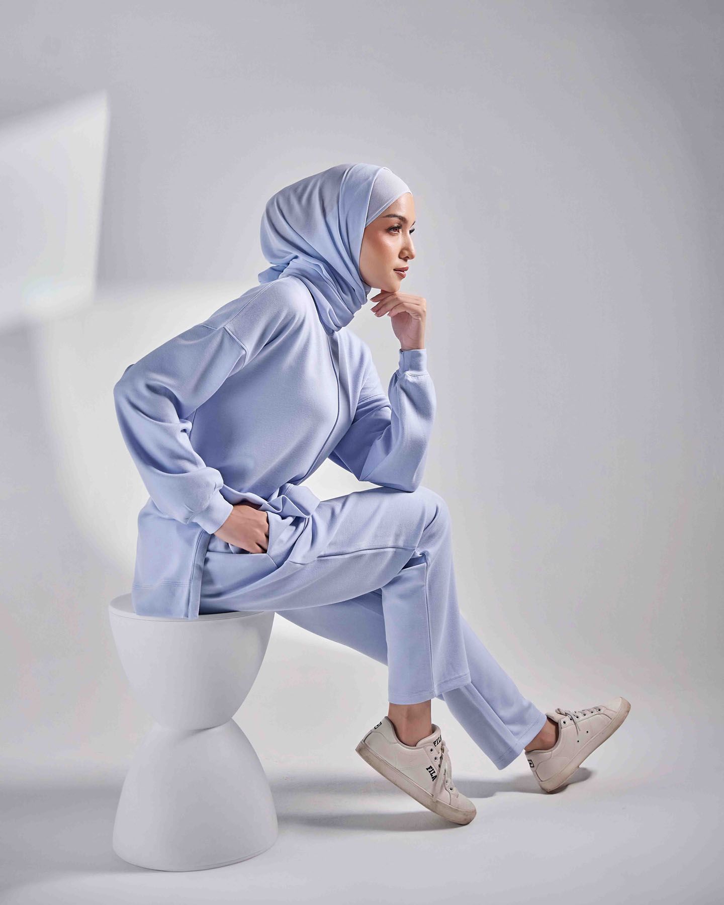 Woman in light blue hijab matching tracksuit seated on white pedestal with white sneakers.