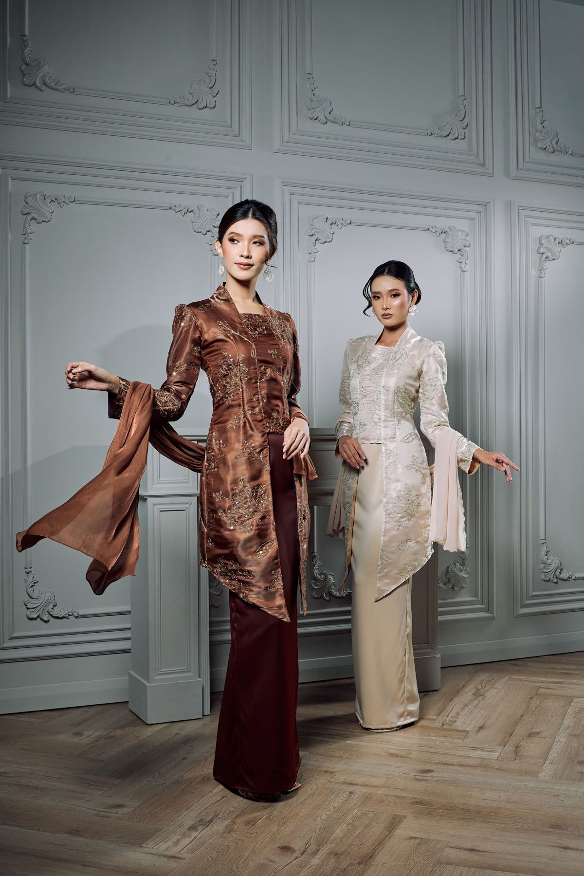 Two models wearing elegant brown and cream embroidered kebaya outfits in a classic white room.