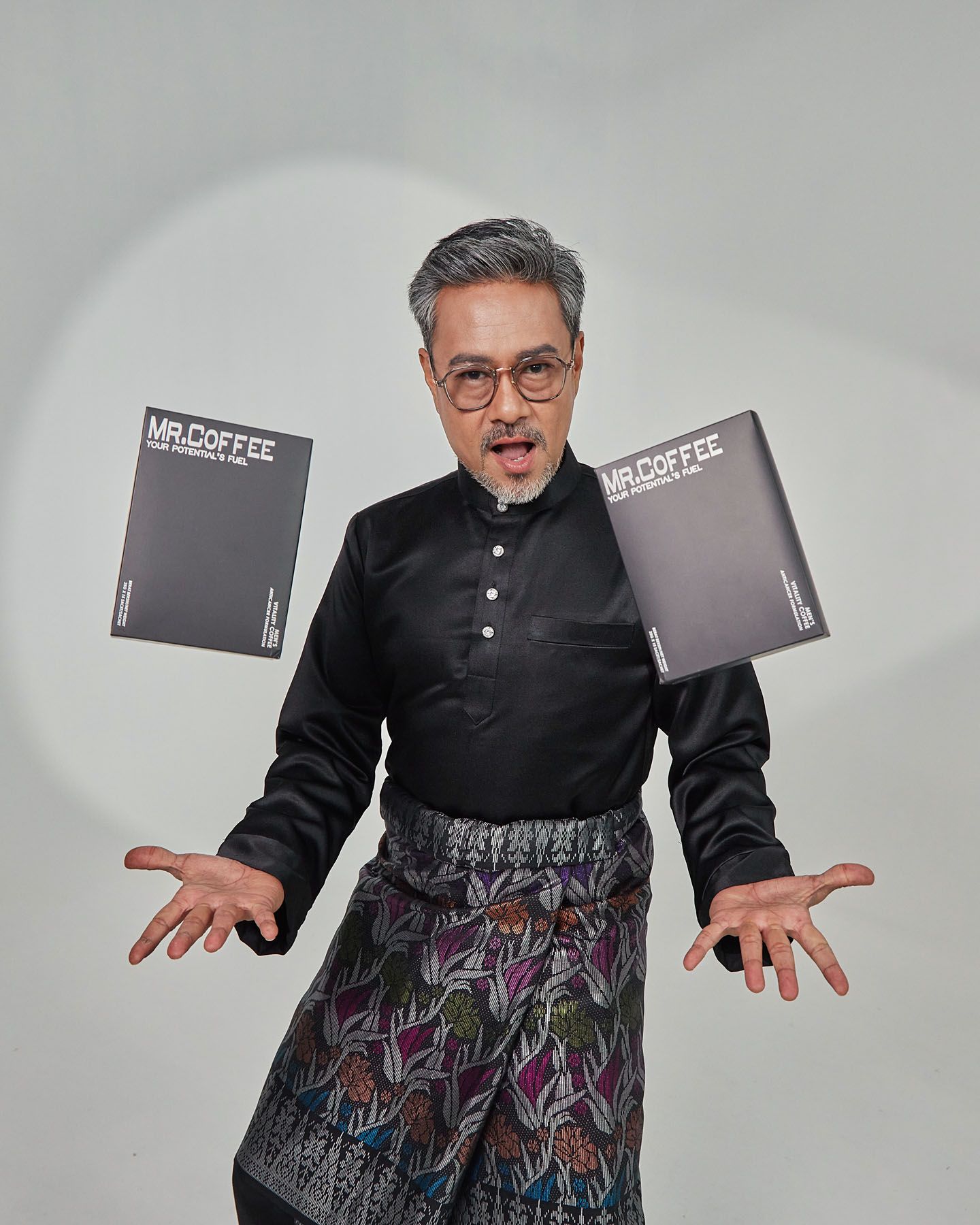 Older man in black baju melayu with colorful sampin tosses books dramatically.