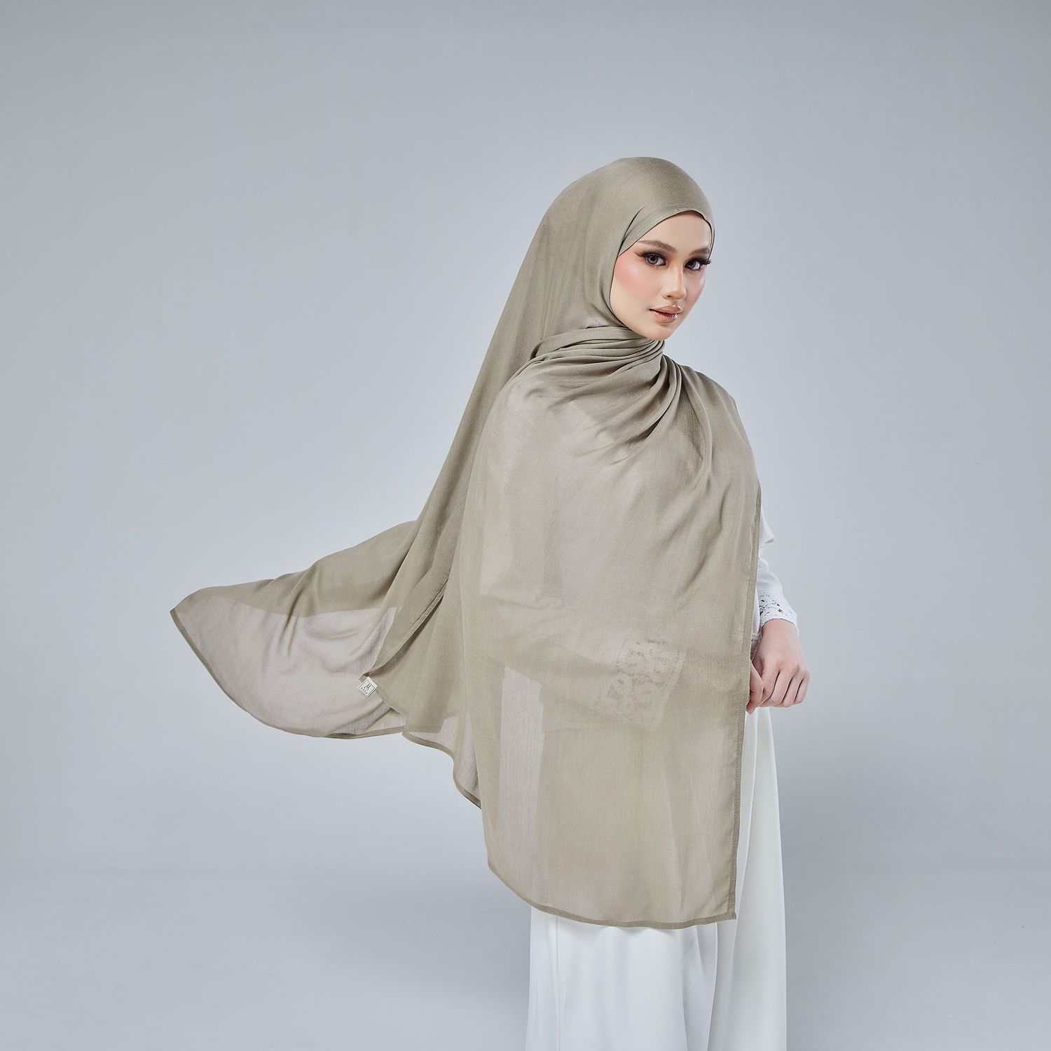 Woman wearing elegant taupe hijab and white dress poses gracefully against neutral background.