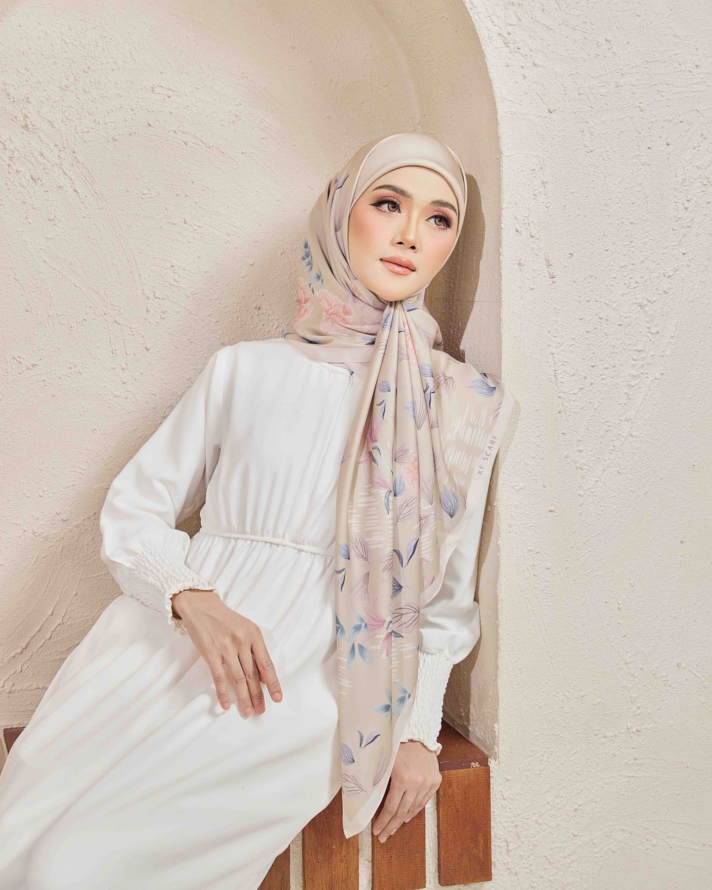 Woman in white dress wearing beige floral printed hijab posed in minimalist architectural setting.