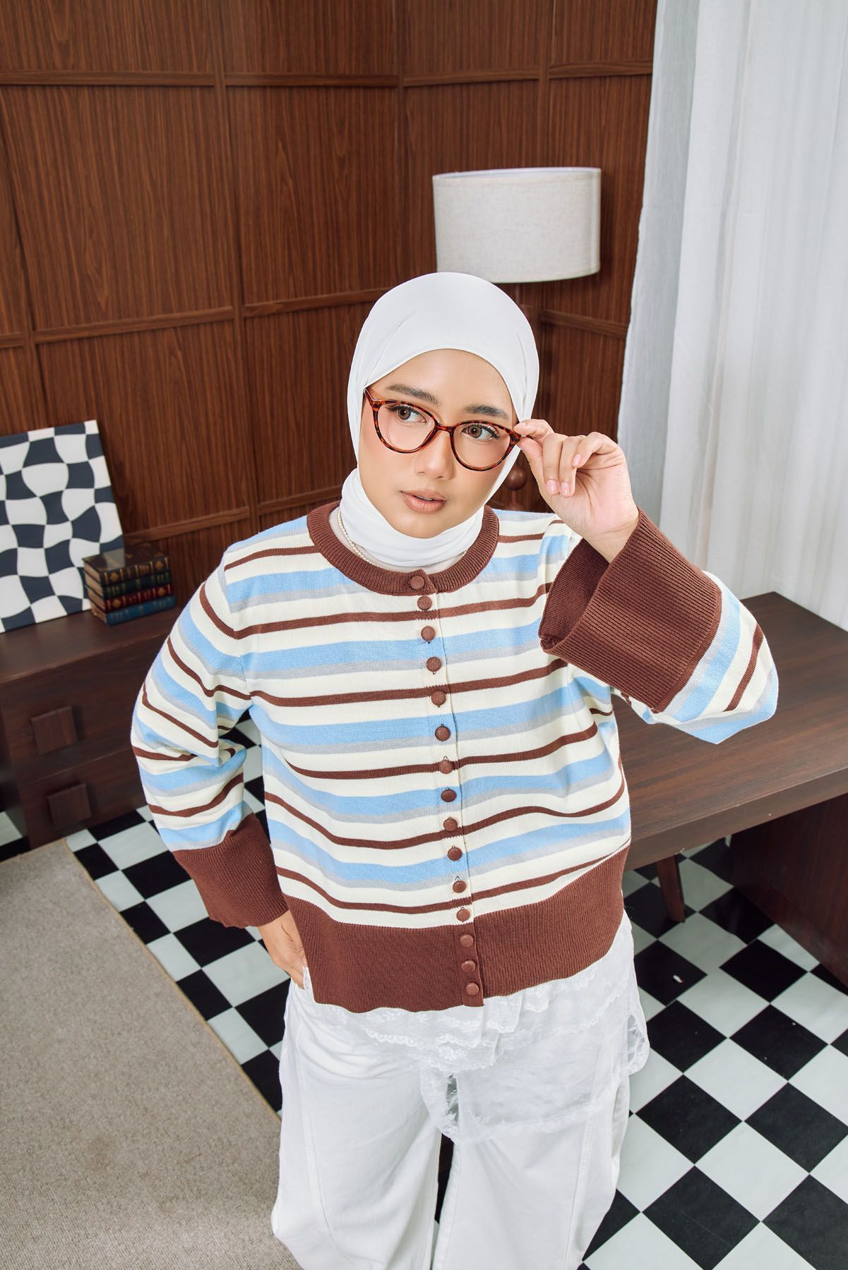 Hijab-wearing woman in striped cardigan adjusts glasses while standing in a retro-styled room with checkered floor.