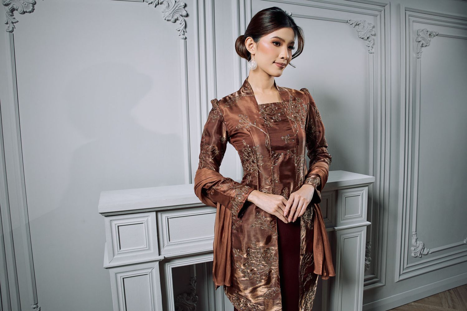 Woman in ornate brown batik kebaya with elegant updo beside white fireplace mantel.