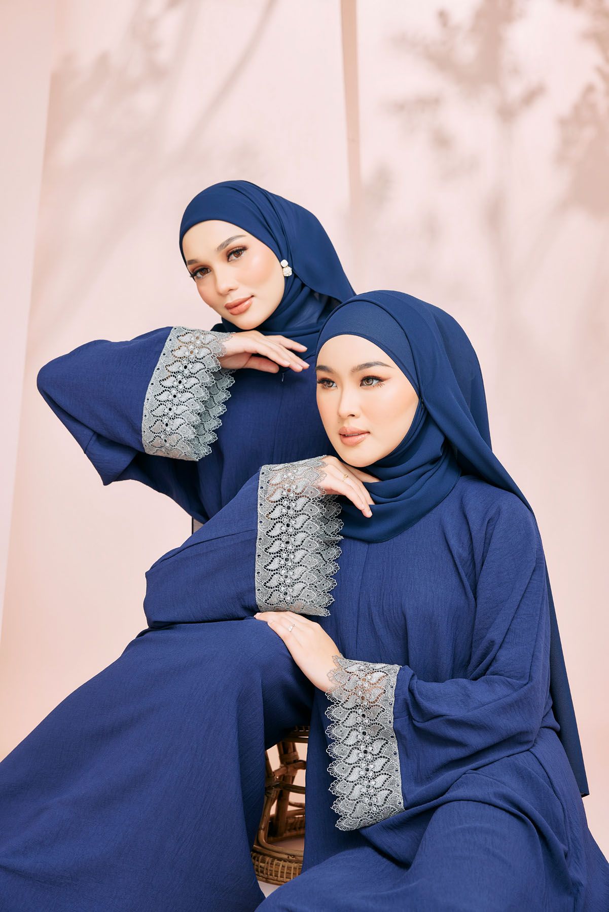 Two women in matching navy blue hijab and abaya with lace cuffs pose elegantly together.