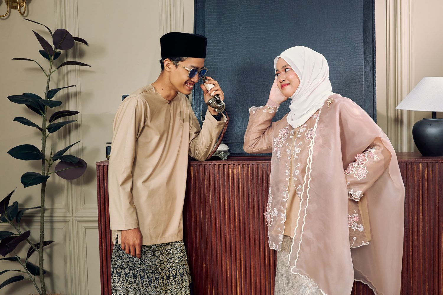 Young Malaysian couple in matching beige traditional Malay attire sharing a playful moment indoors.