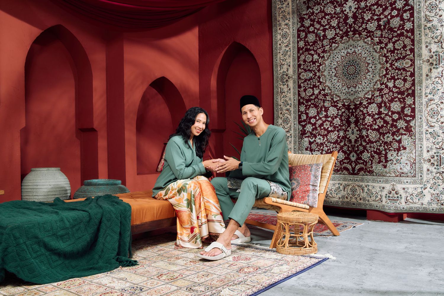 Malay couple in matching green baju kurung and baju melayu sitting together in a Moroccan-inspired red studio.