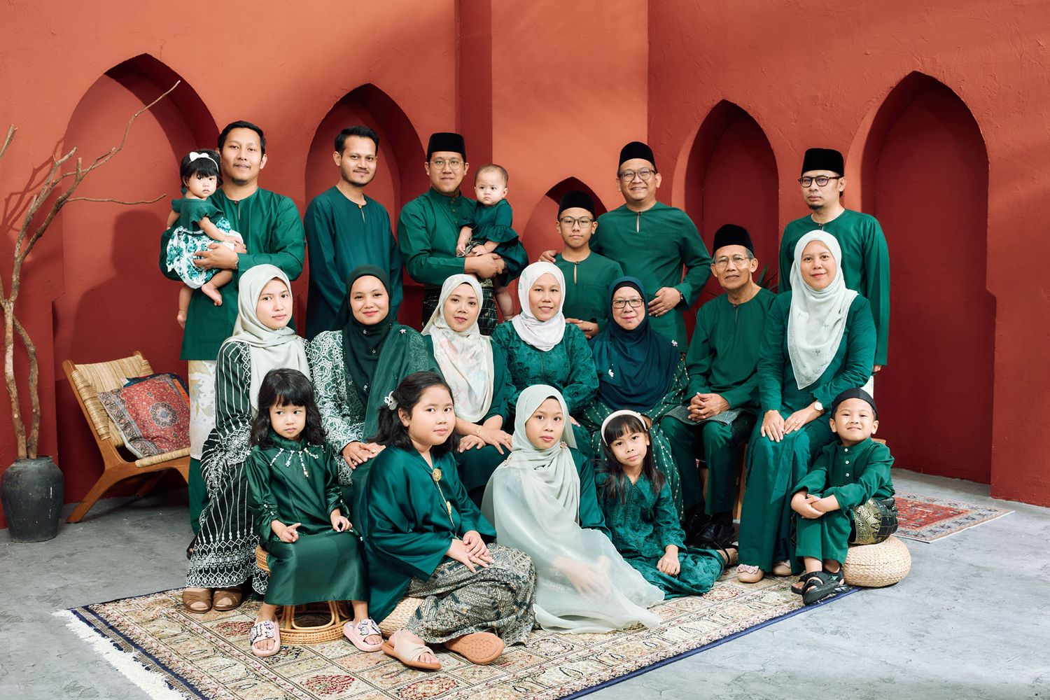 Large Malaysian family in coordinating green traditional Hari Raya outfits posing together in a red studio.
