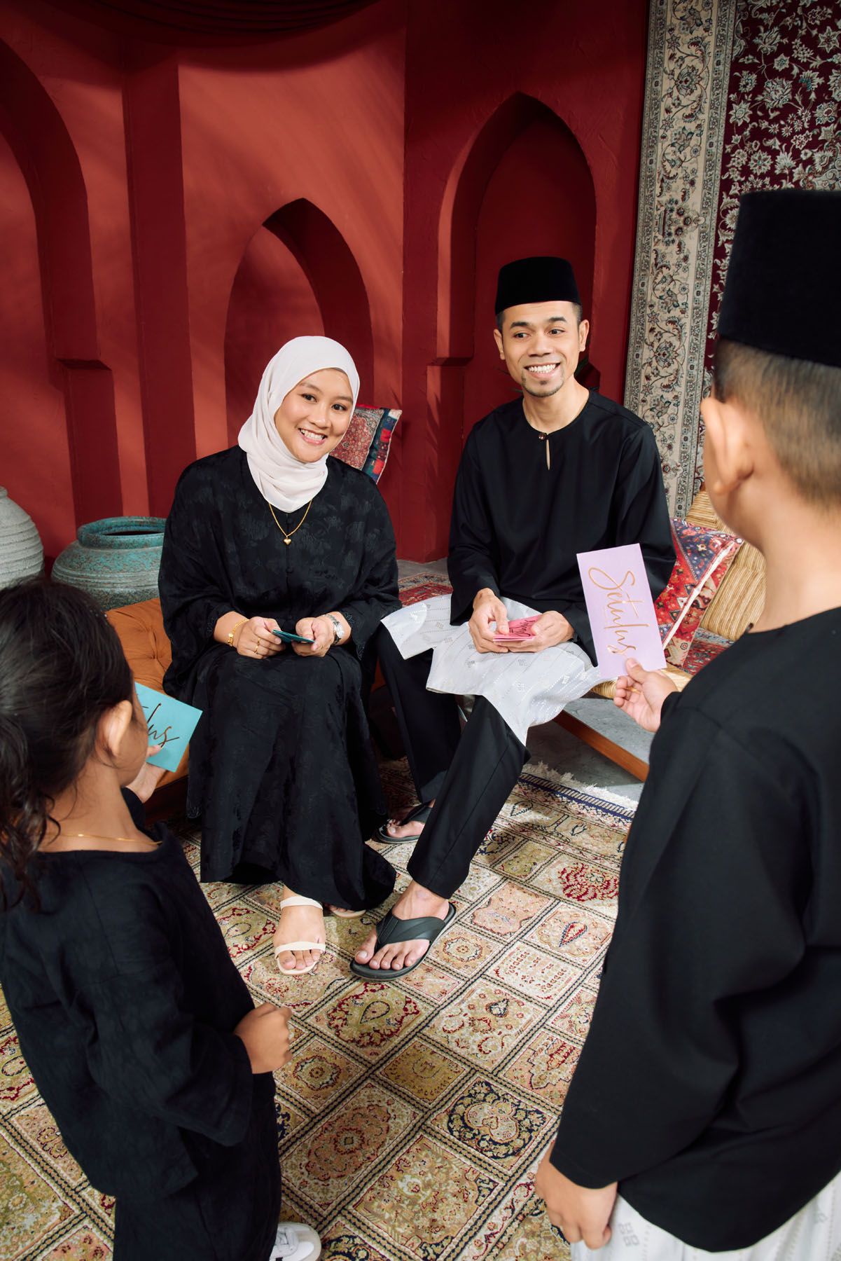 Muslim couple in black traditional attire giving Hari Raya green packets to children.