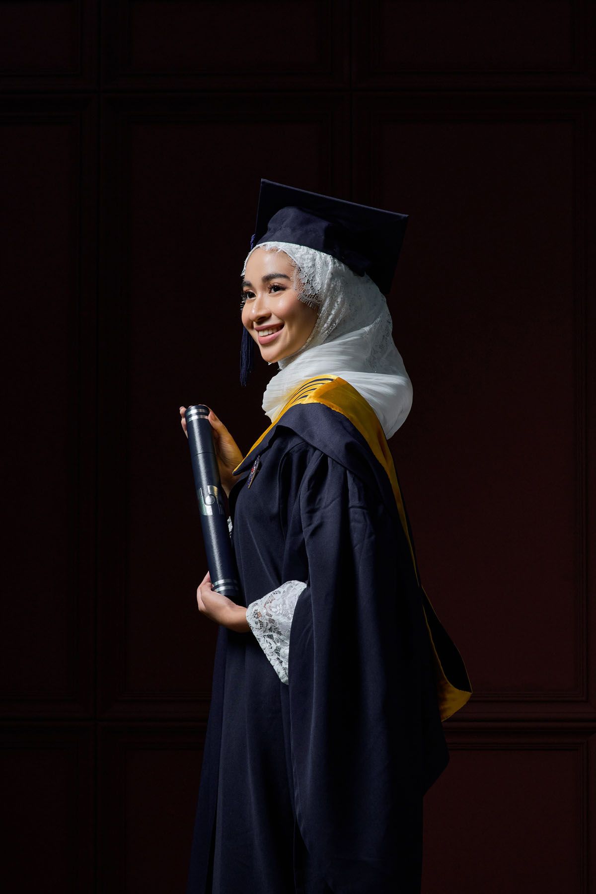 Smiling Muslim woman in navy graduation gown, hijab, and mortarboard cap holding diploma scroll.