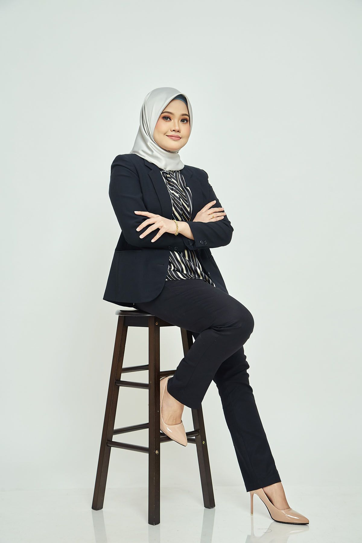 Young Muslim woman in black blazer and white hijab sitting on stool with arms crossed gracefully.