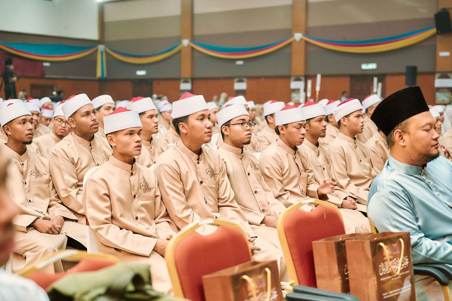 Rows of young male students in matching beige uniforms and white caps attending an Islamic Quran ceremony.