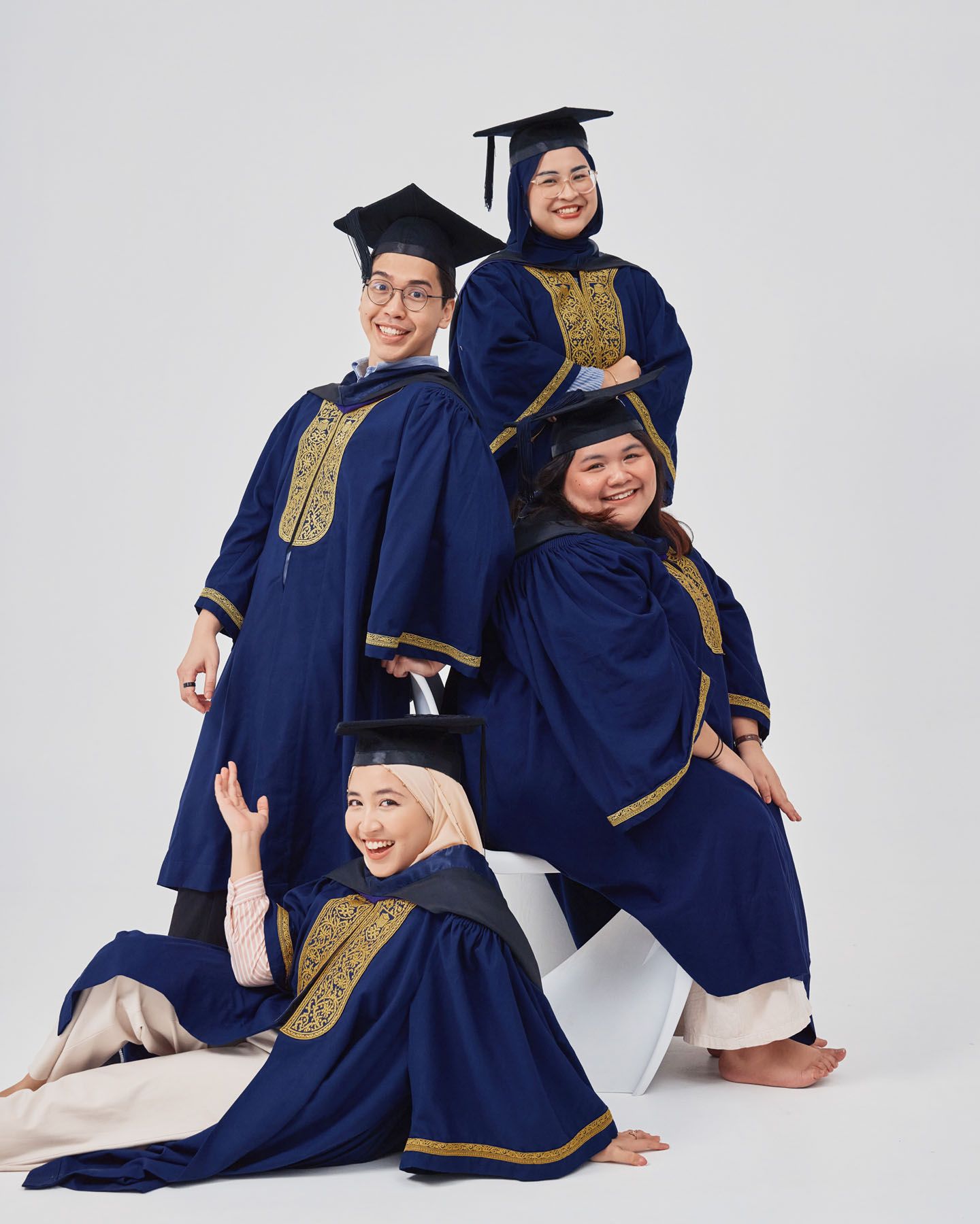 Four graduates in navy and gold robes posing playfully on white props for graduation photos.