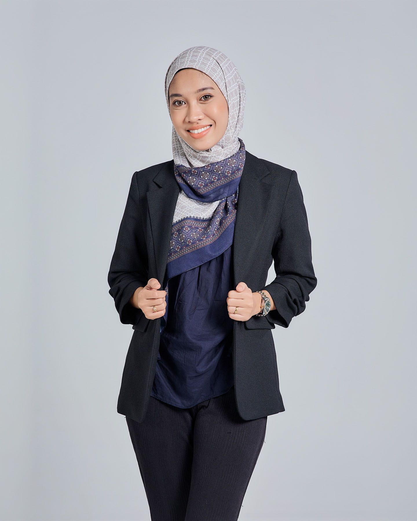 Smiling Muslim woman in black blazer and patterned hijab posing casually on gray background.