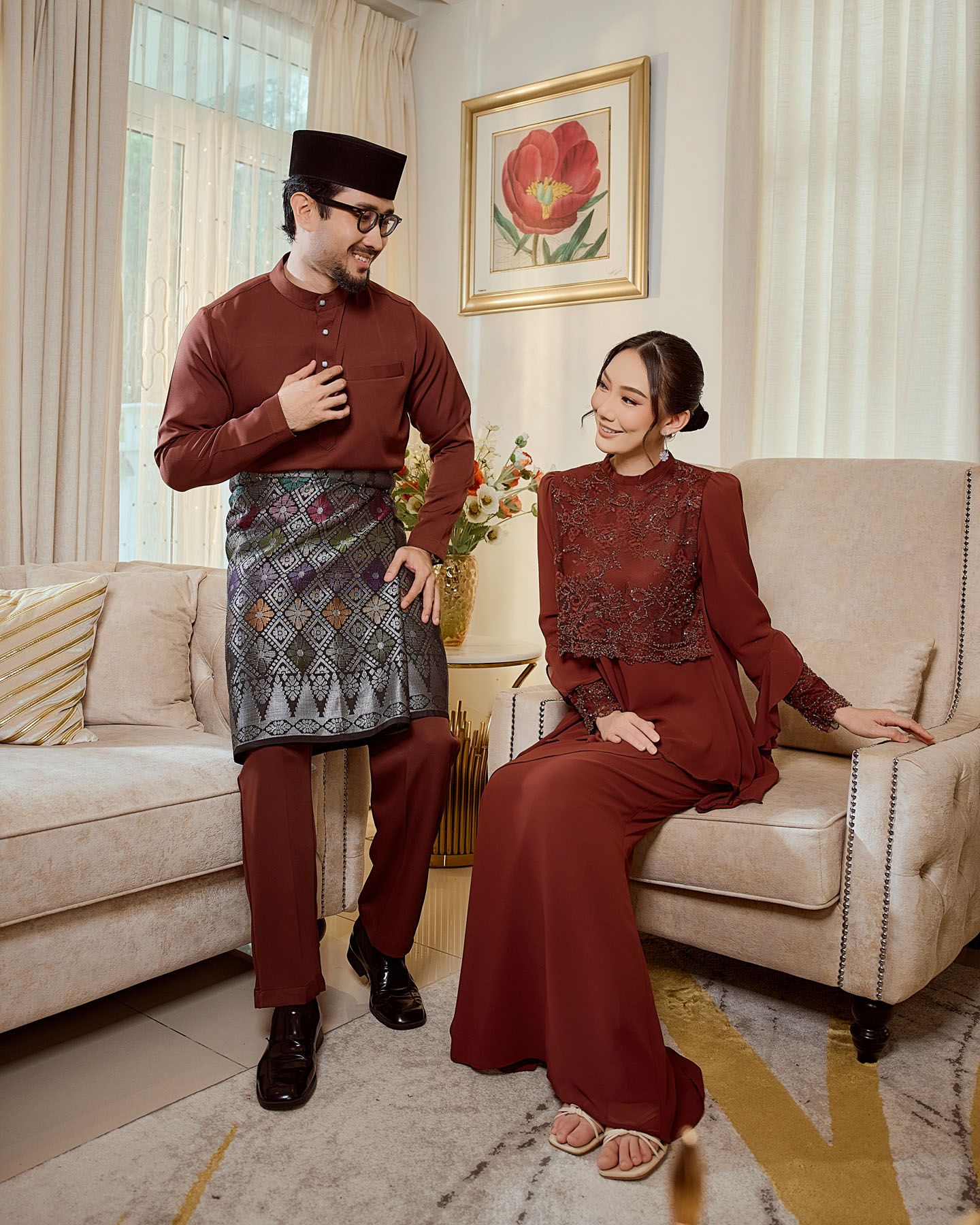 Couple in matching maroon traditional Malaysian outfits posing in an elegant living room setting.