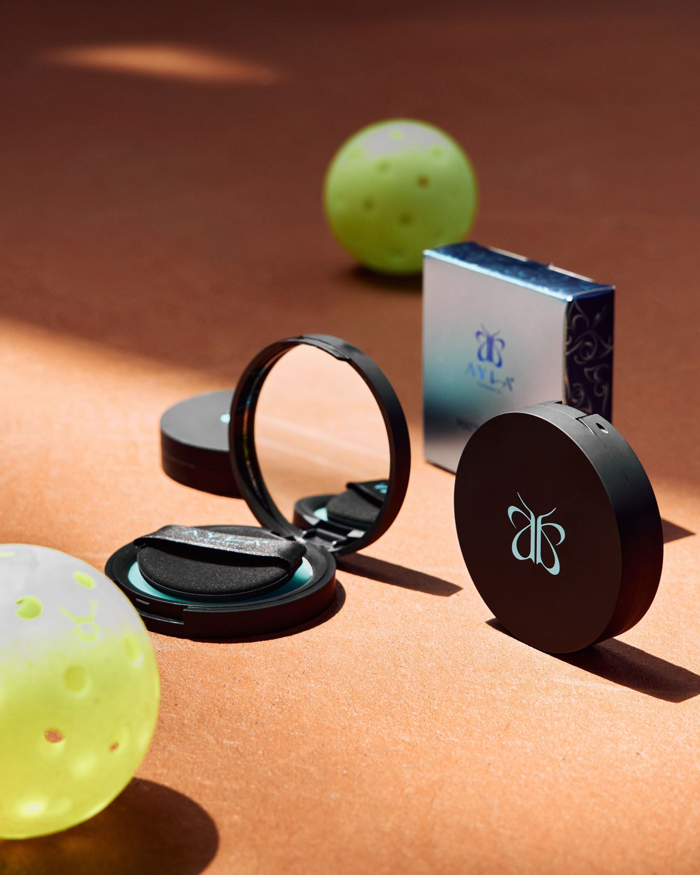 Open black compact powder with butterfly logo beside yellow pickleball balls on warm terracotta surface.