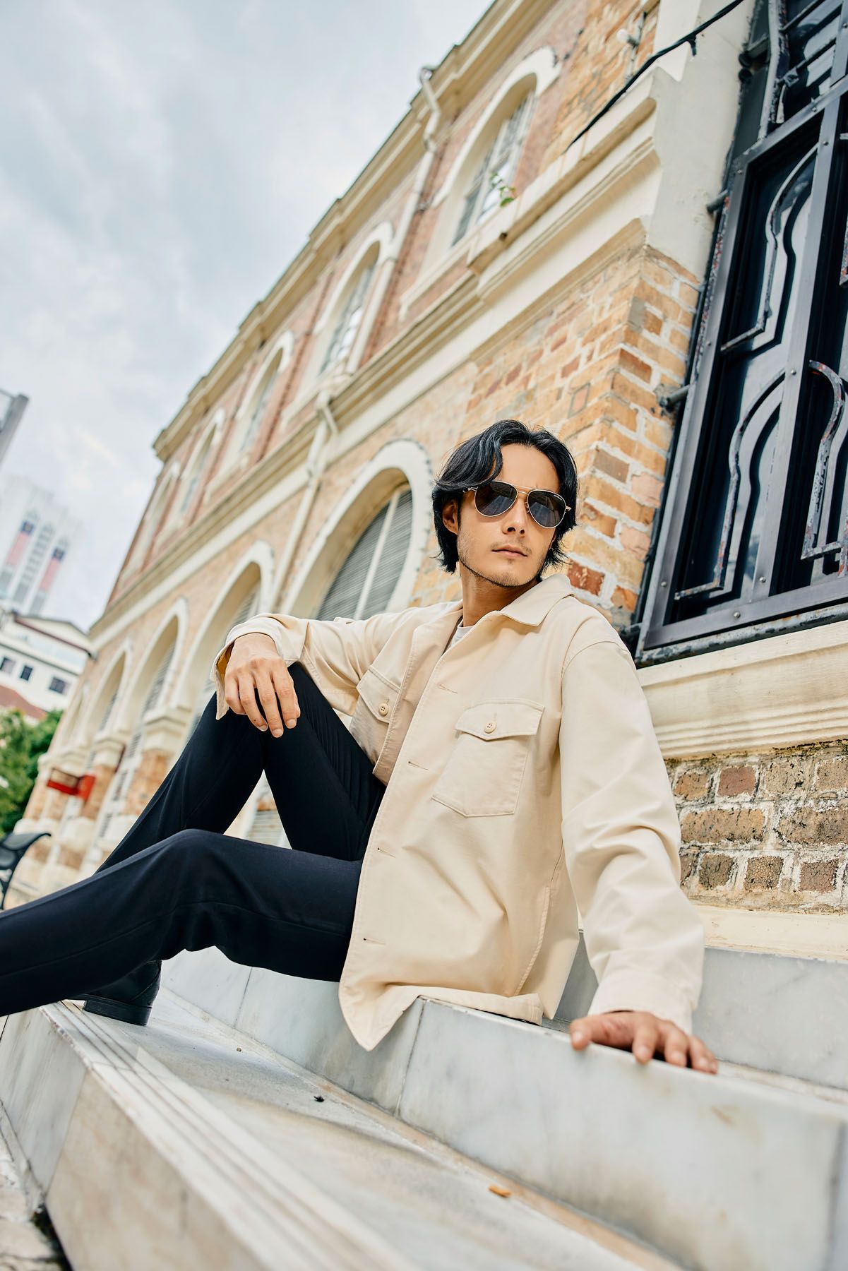 Stylish man in beige overshirt and black pants wearing sunglasses on outdoor steps.