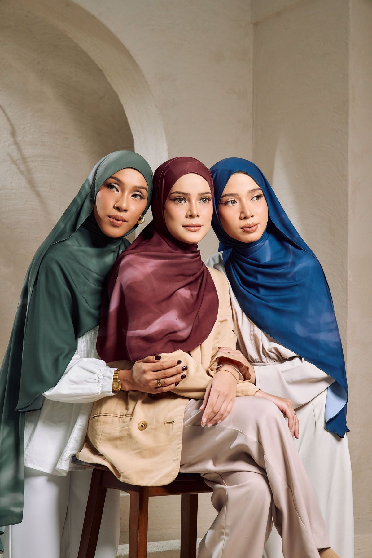 Three women wearing colorful hijabs in green, burgundy, and blue pose together in an elegant architectural setting.
