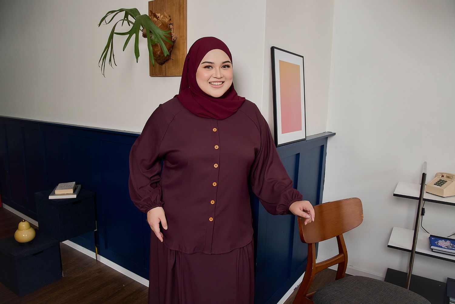 Plus-size Muslim woman smiling in maroon hijab and matching button-up dress in modern interior setting.