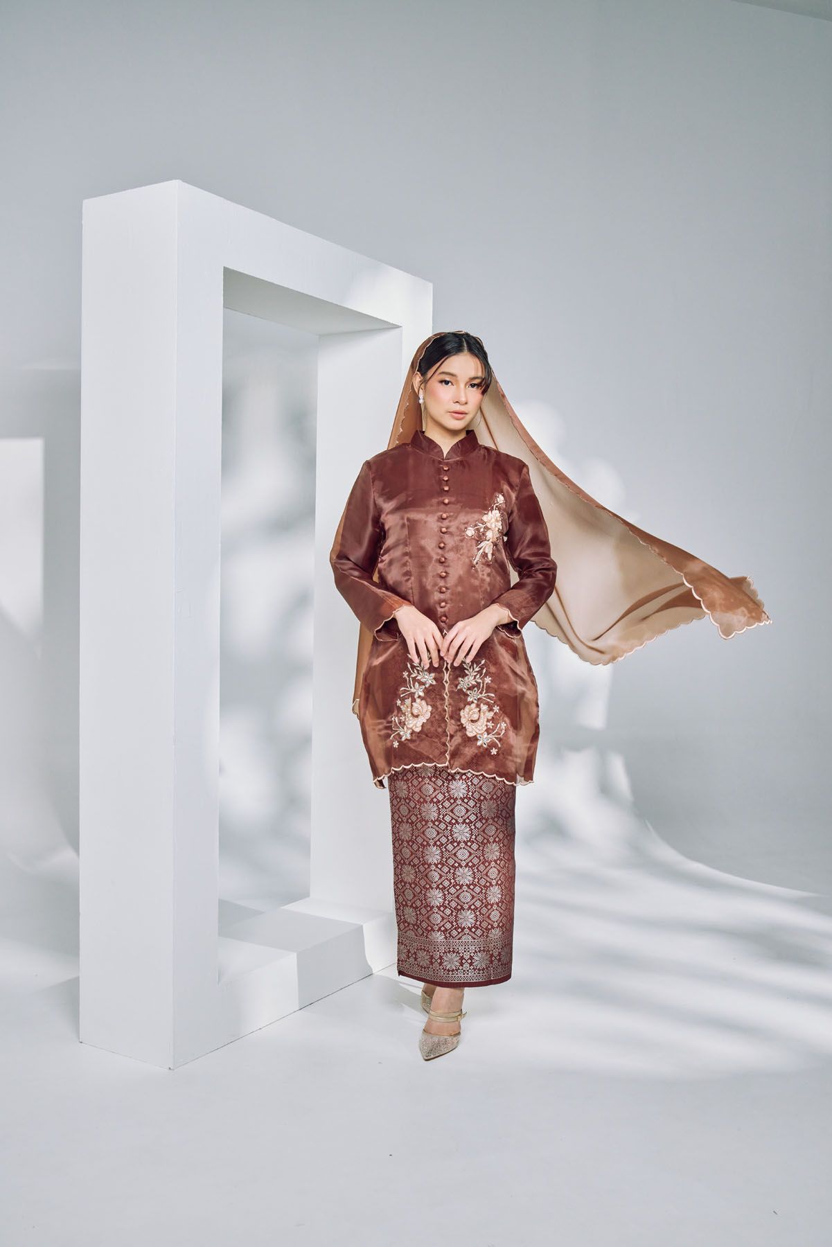 Woman in brown embroidered kebaya with batik skirt and flowing shawl posing in a white studio.