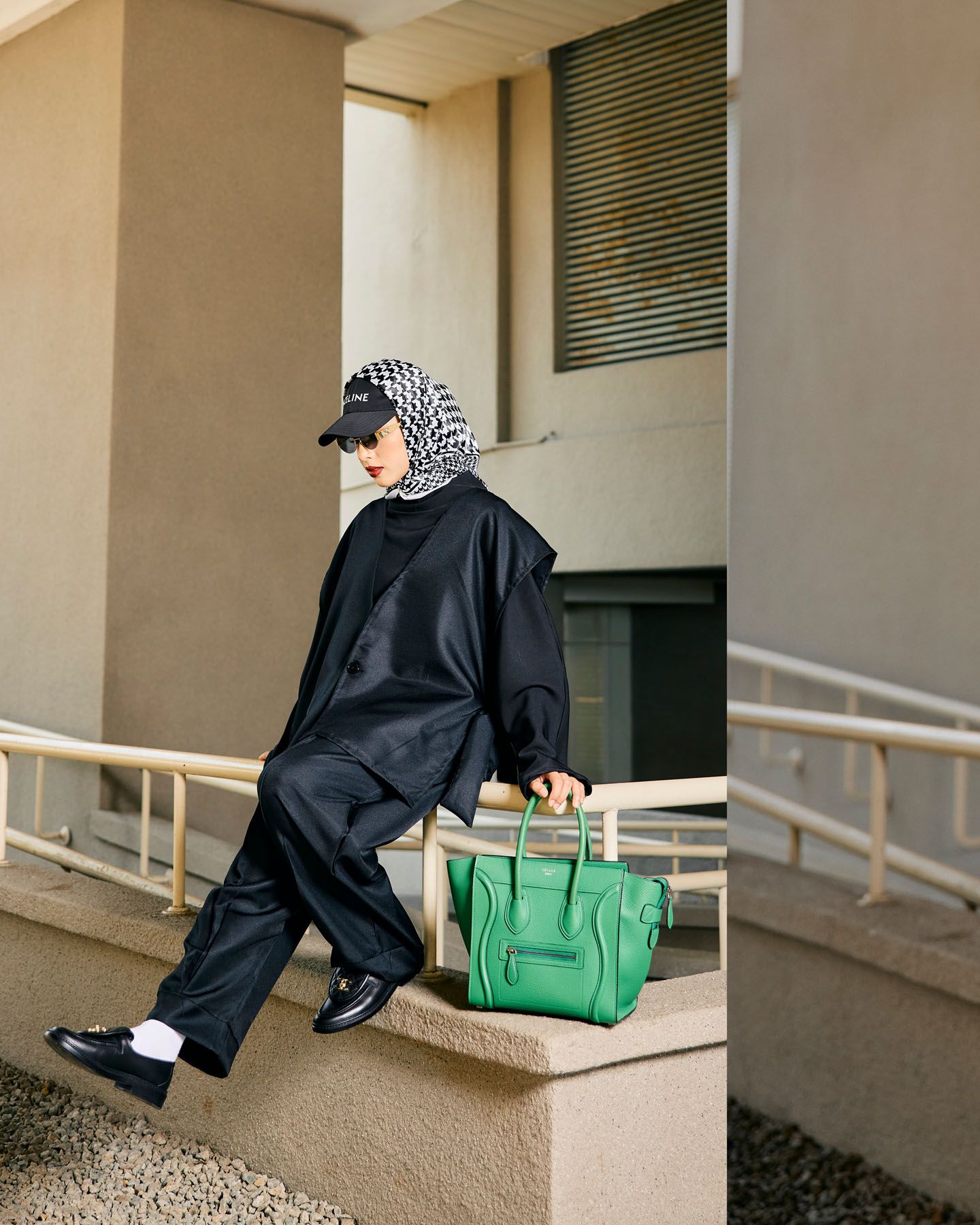 Stylish woman in black outfit and patterned hijab carrying green Celine bag on outdoor steps.