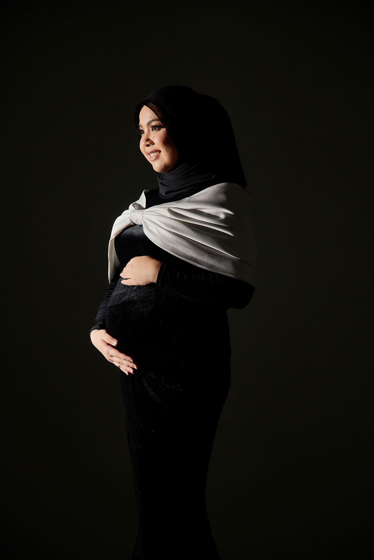 Pregnant Muslim woman in black hijab and dress cradling her baby bump against a dark studio background.