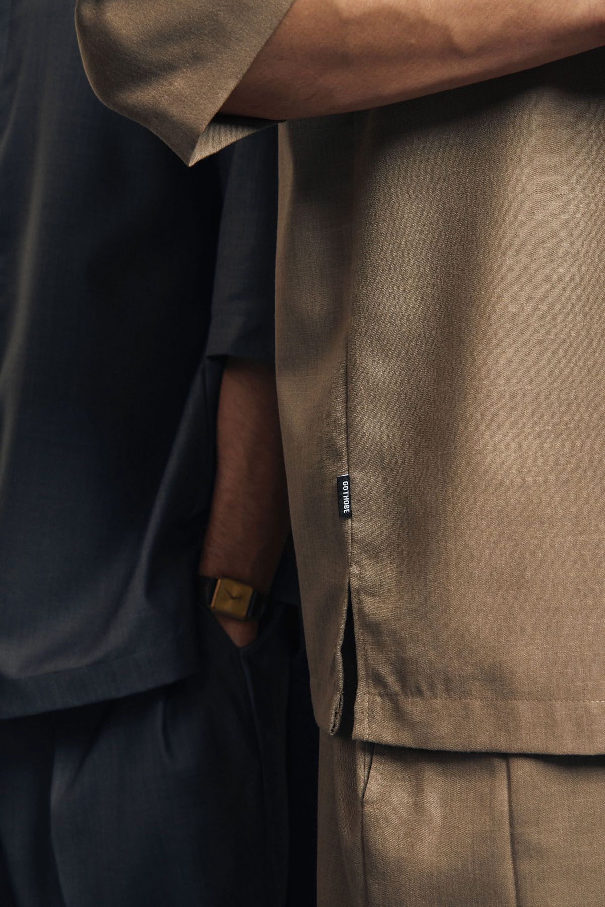 Close-up detail of a beige linen shirt hem with small brand label next to dark fabric.