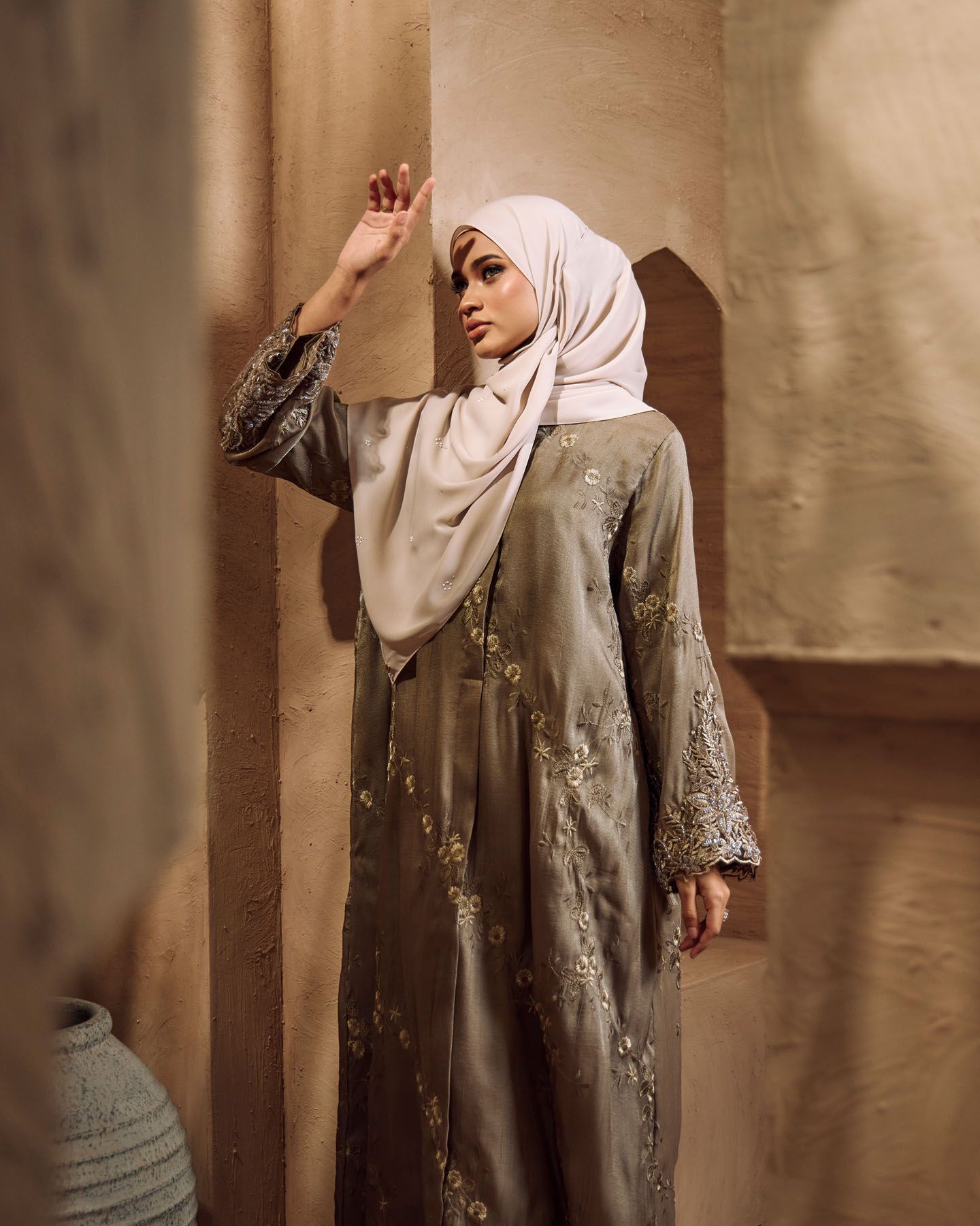 Woman in olive embroidered abaya and cream hijab leans against wall.