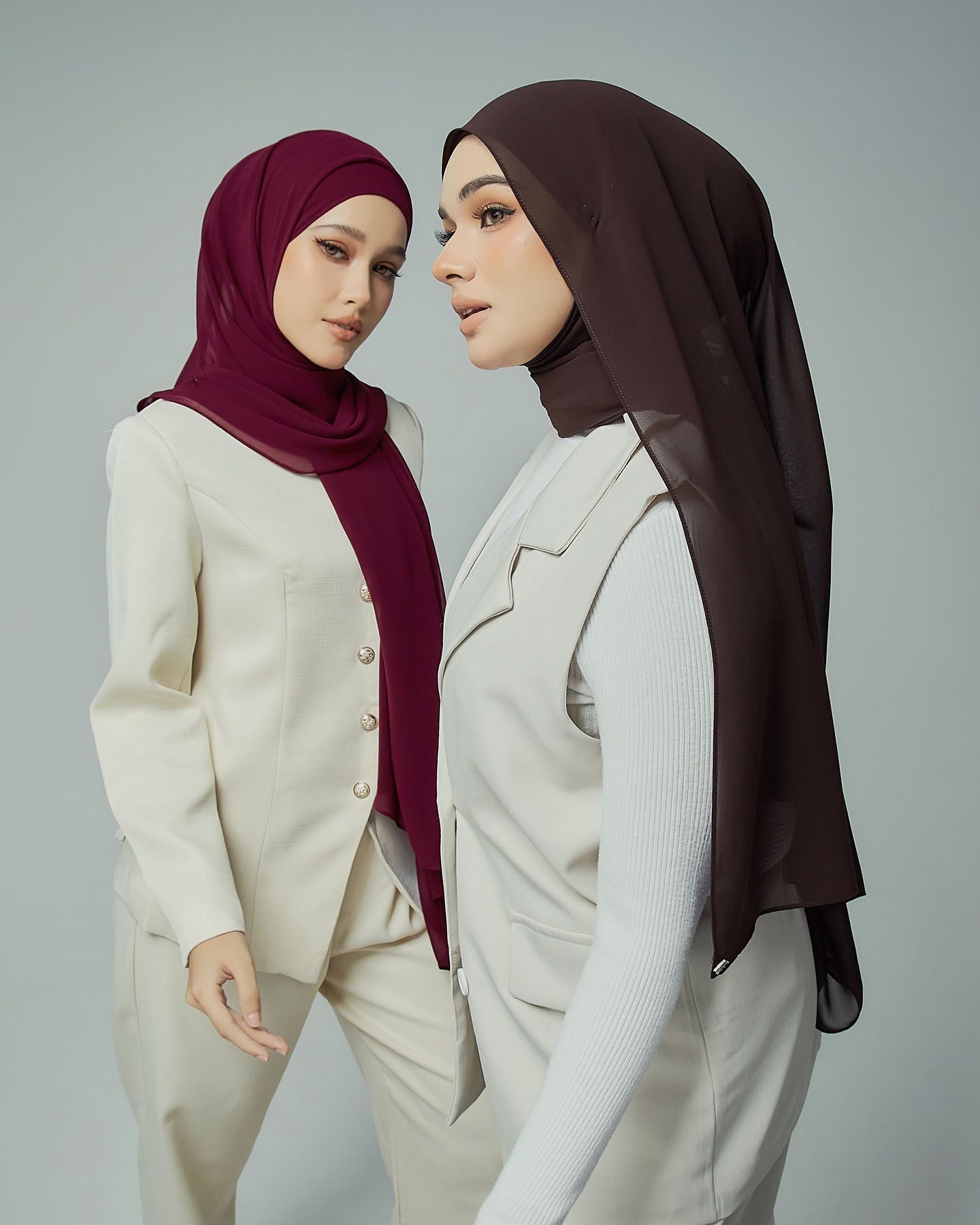 Two women in burgundy and dark brown chiffon hijabs with white tailored suits in studio portrait.