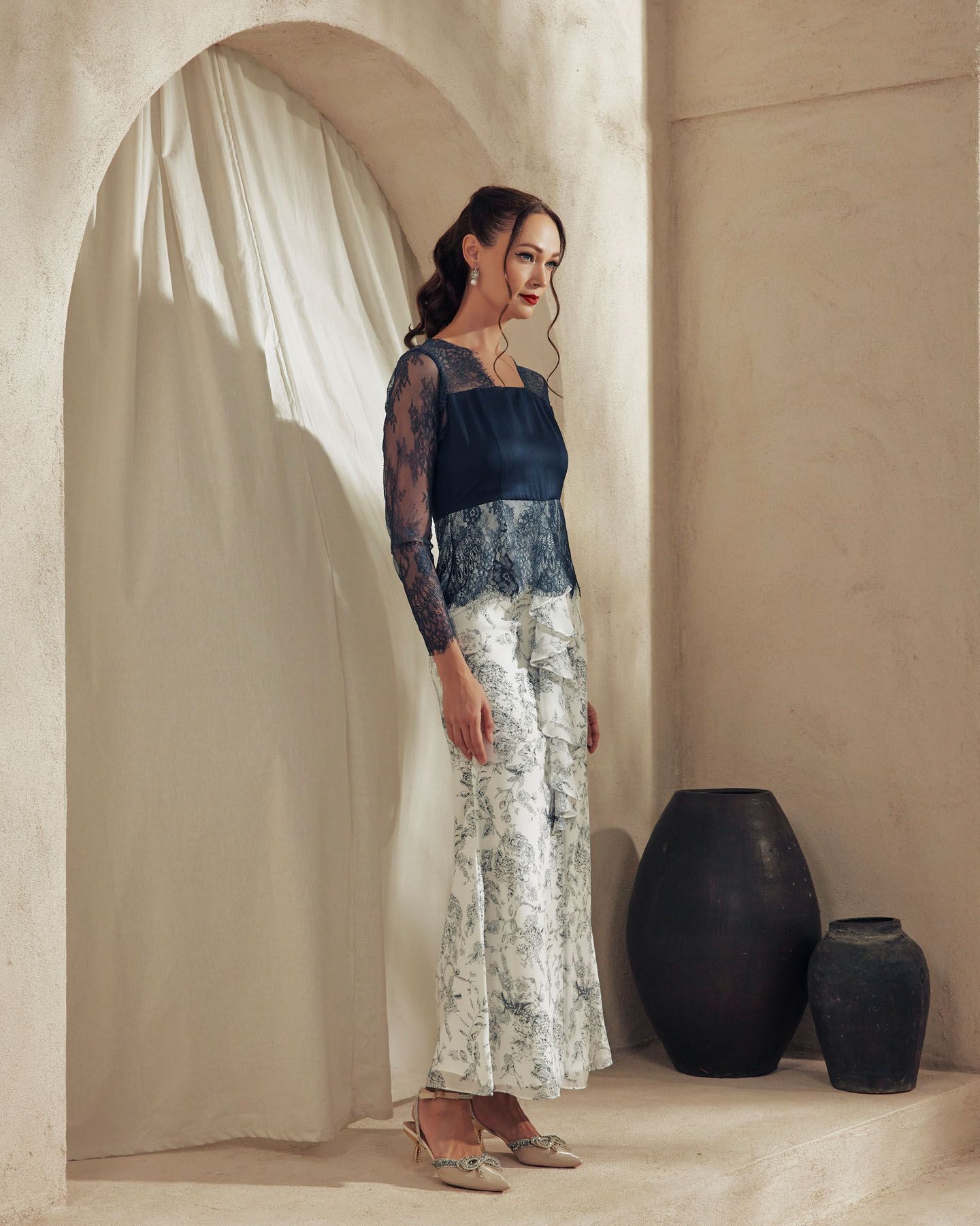 Woman in navy lace top and floral wide-leg pants poses in minimalist archway with black vases.