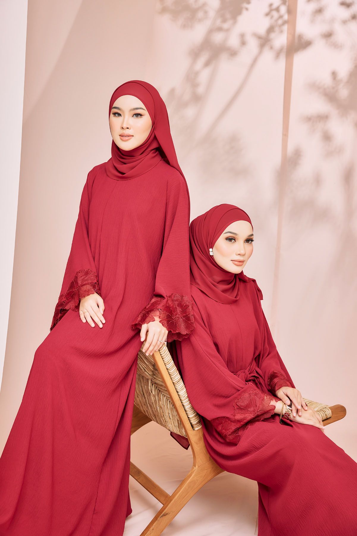 Two women in matching red hijab and abaya with lace cuffs pose in a warm-toned studio.