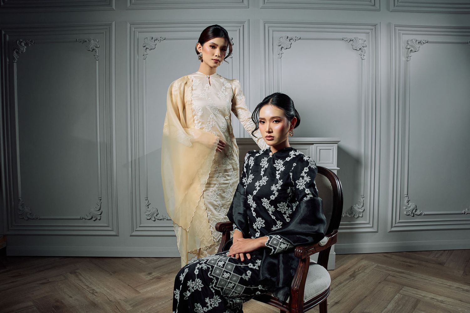 Two women in traditional attire, one in black floral kebaya seated and one standing in cream outfit, elegant interior.