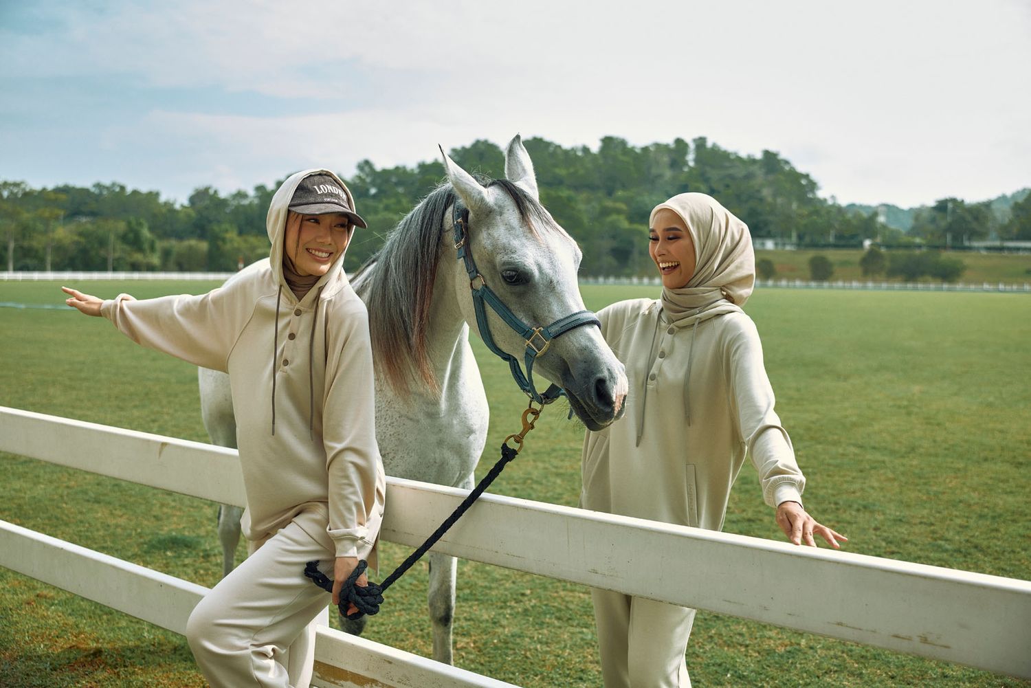 Two women in cream modest outfits smiling beside white horse at equestrian fence.