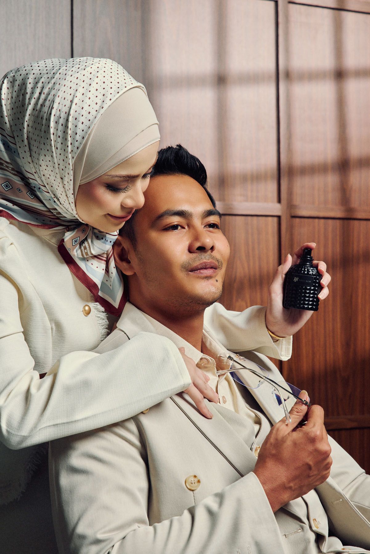 A couple in matching beige outfits examine a dark perfume bottle together indoors.