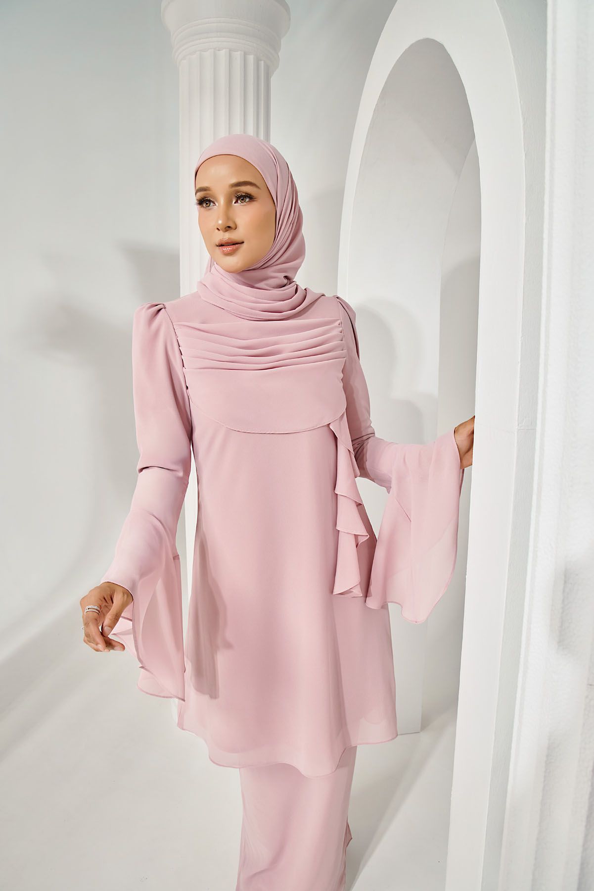 Woman in soft pink modest tunic with bell sleeves and matching hijab, posing near white arched columns.