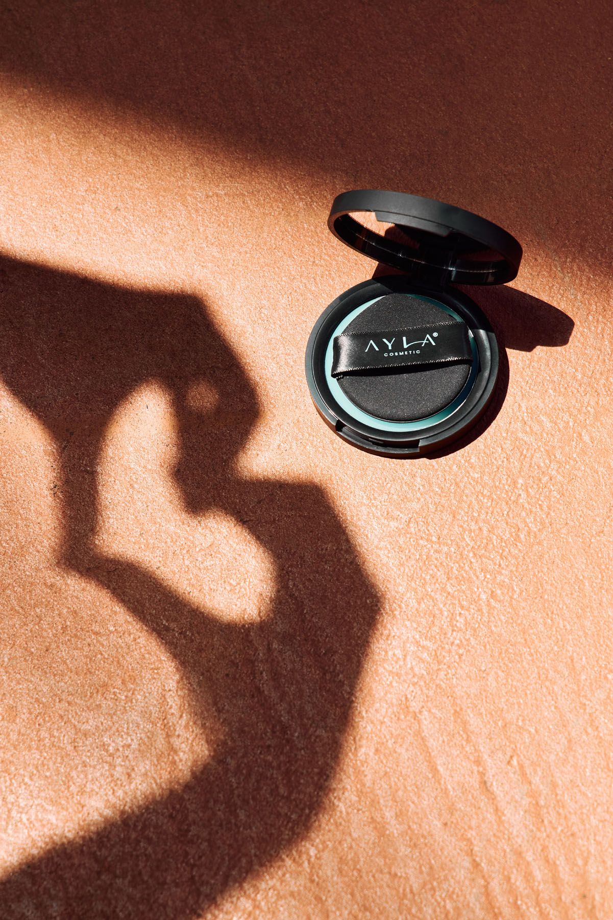 AYLA cosmetics black compact powder on clay surface with dramatic hand shadow in bright sunlight.