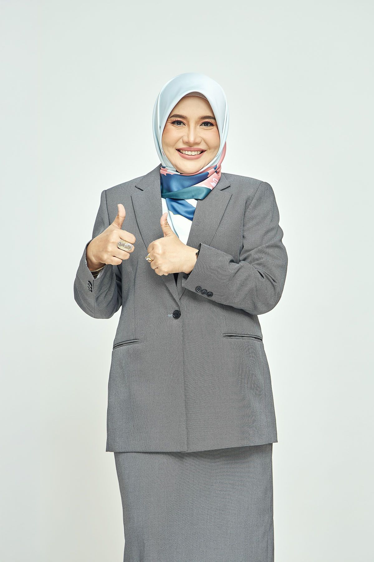 Smiling Muslim businesswoman in gray blazer and hijab giving two thumbs up gesture.