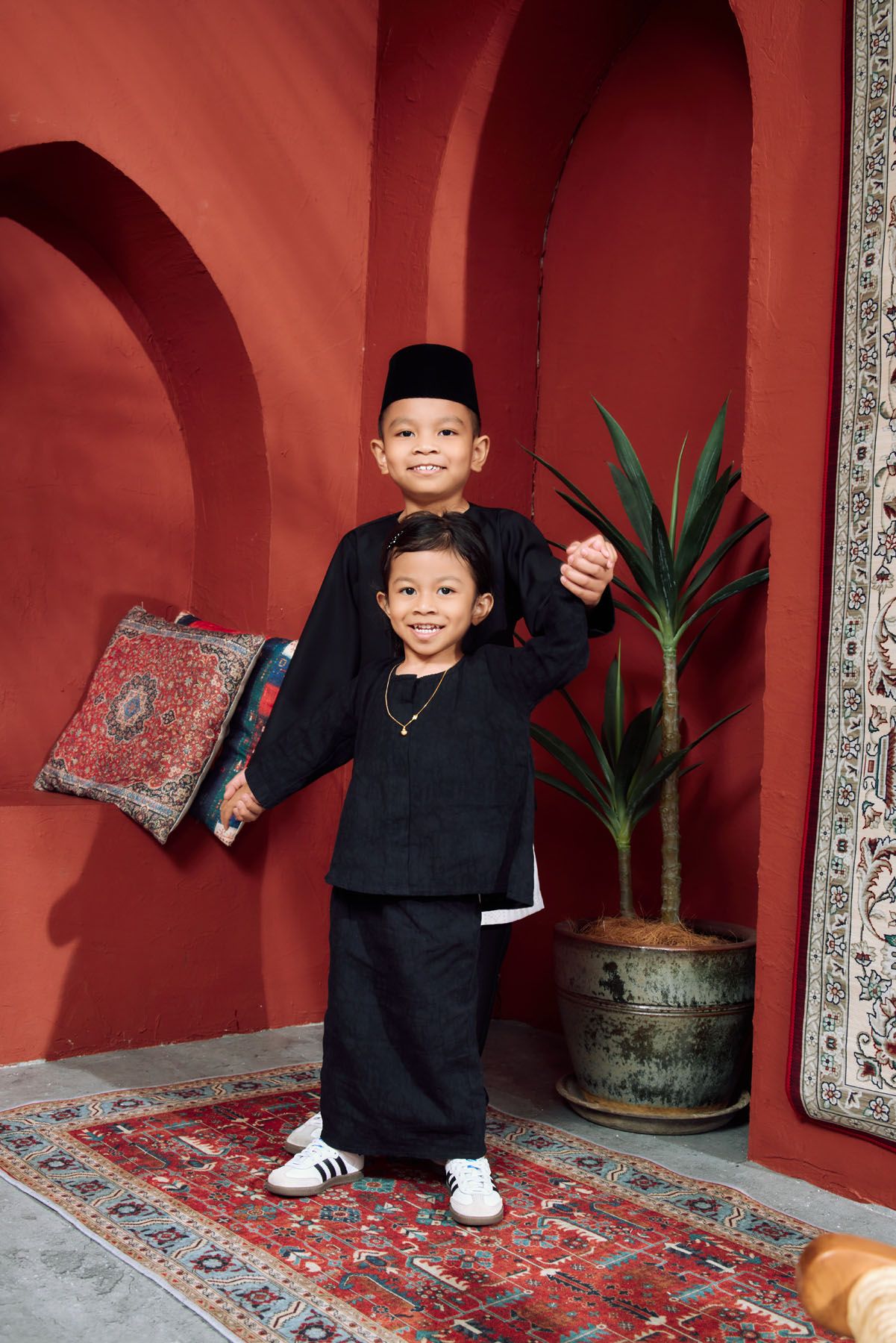 Two young Malaysian boys in matching black Baju Melayu traditional outfits posing against red wall.