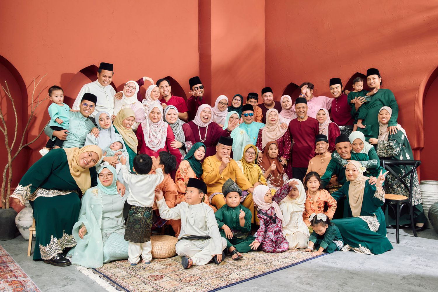 Large Malaysian family group photo in traditional Eid Baju Melayu and Baju Kurung attire in green, pink, and earth tones.
