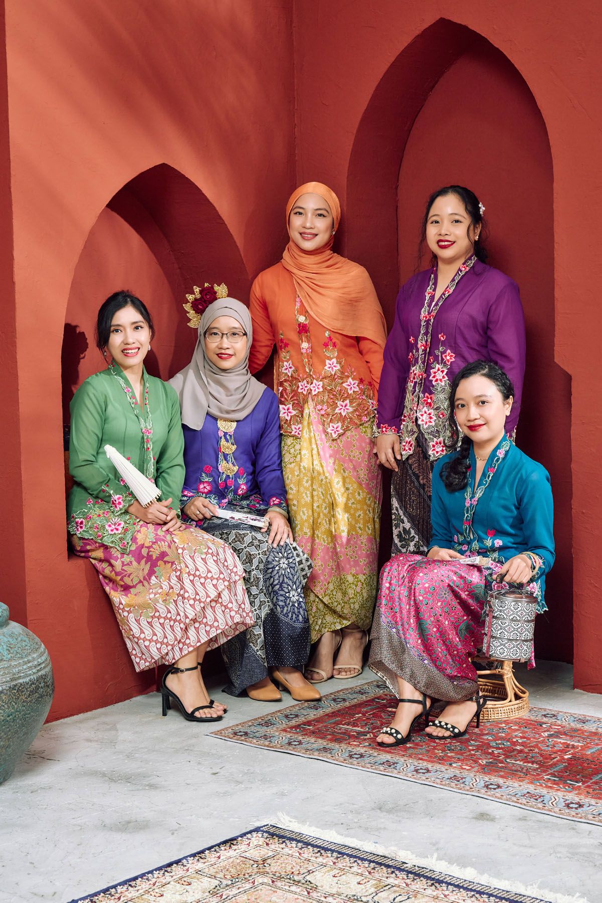 Group of five women wearing vibrant traditional Malay kebaya dresses posing in front of red arched backdrop.