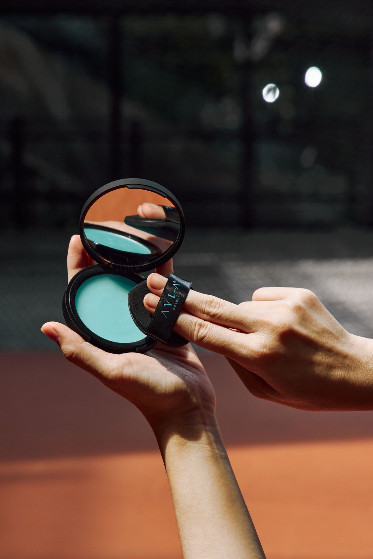 Hands holding open AYLA black compact with teal powder and mirror reflecting skin on dark court background.