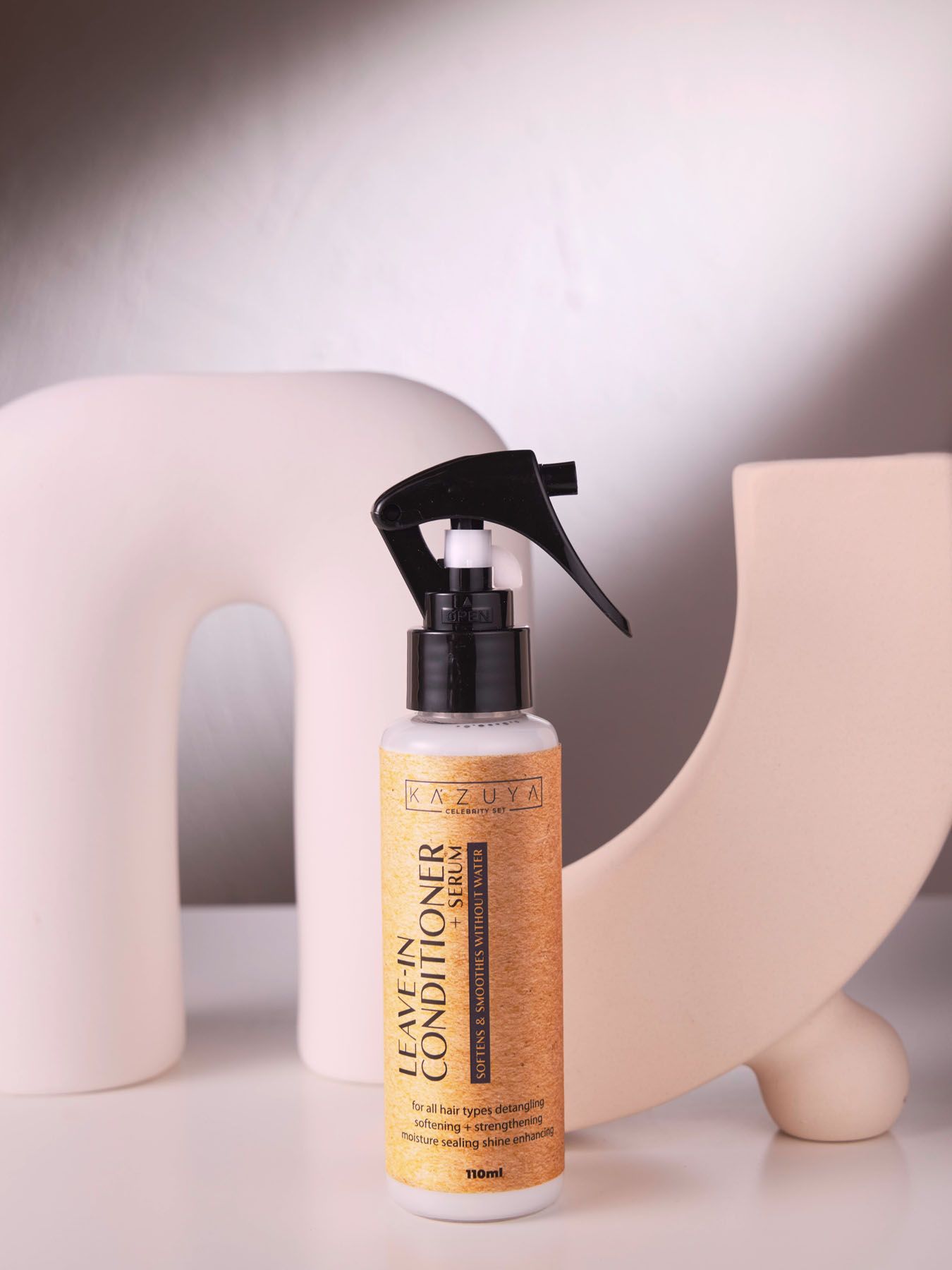 KAZUYA Leave-In Conditioner Spray 150ml golden bottle with trigger sprayer for all hair types on beige backdrop.