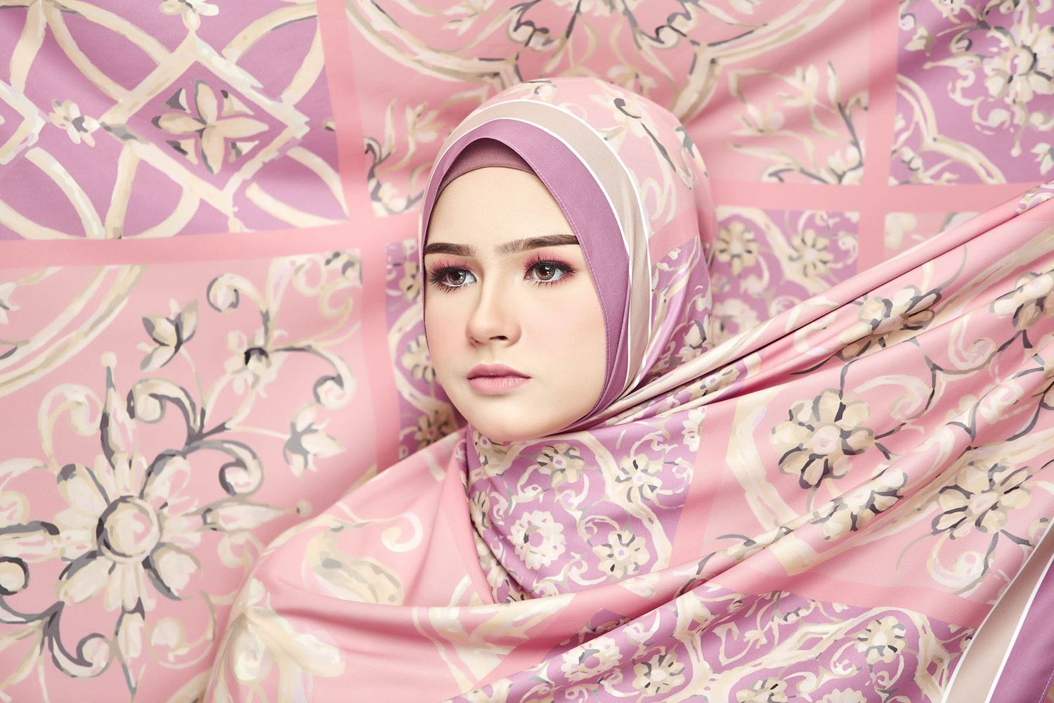 A young woman in a pink floral hijab poses surrounded by a matching pink patterned fabric.