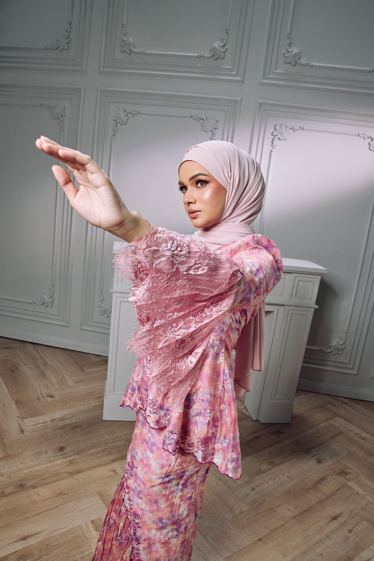 Muslim woman in pink floral kebaya and hijab posing gracefully in white room.