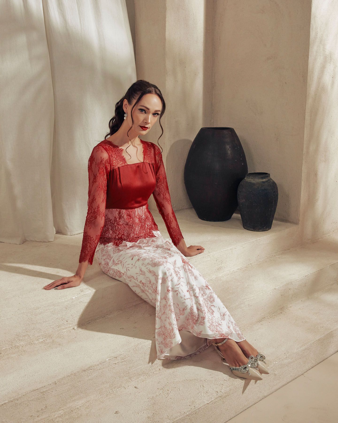 Woman in red lace top and floral skirt sits gracefully in minimalist setting with black ceramic vases.